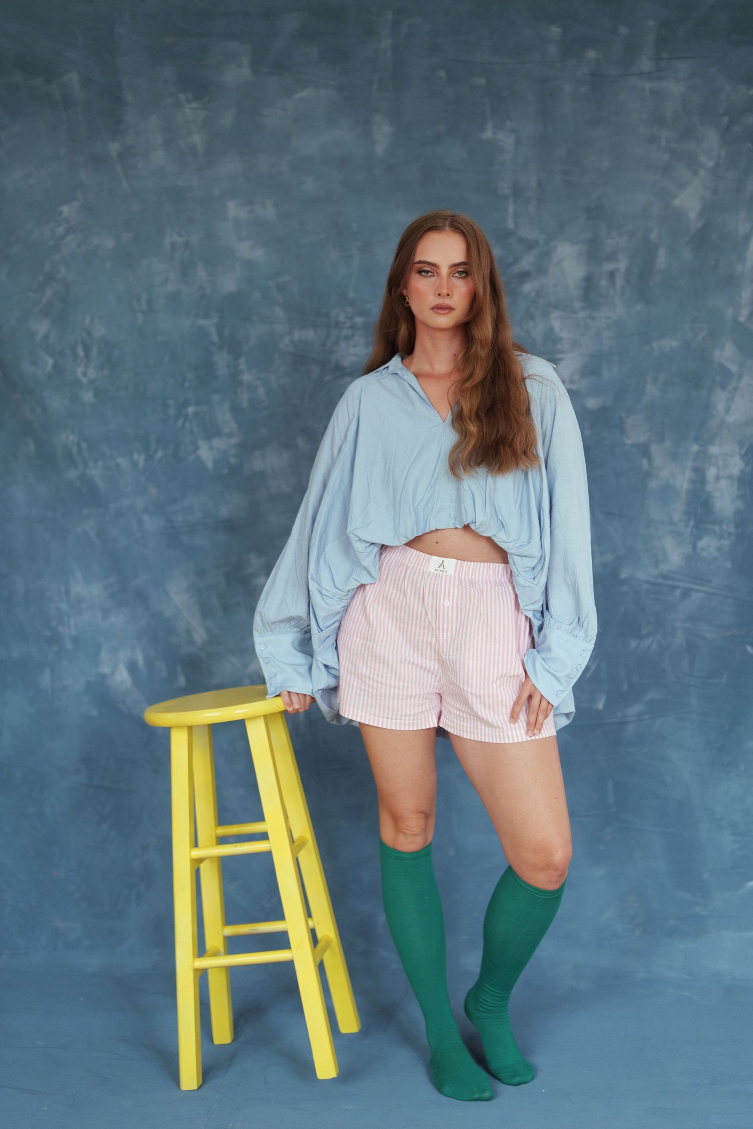Young woman in blue oversized shirt, pink shorts, and green knee socks posing next to a yellow stool on blue background.