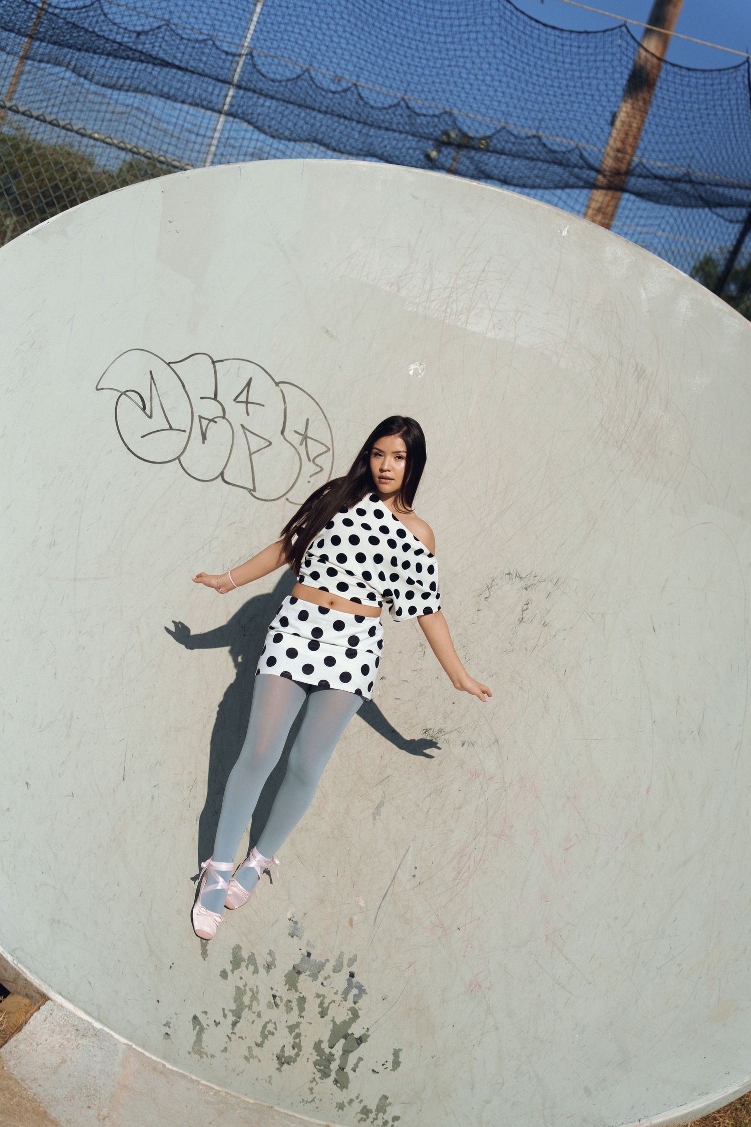 Someone in a polka dot shirt and gray pants performs tricks at a skate park ramp.