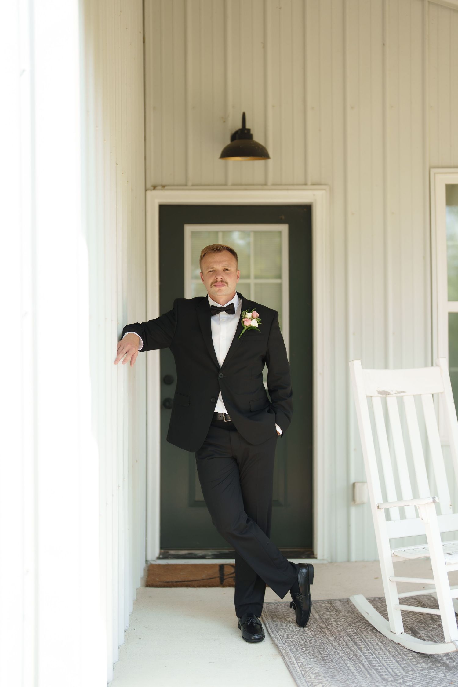 Groom in black tuxedo with boutonniere leaning against white farmhouse porch column beside rocking chair.