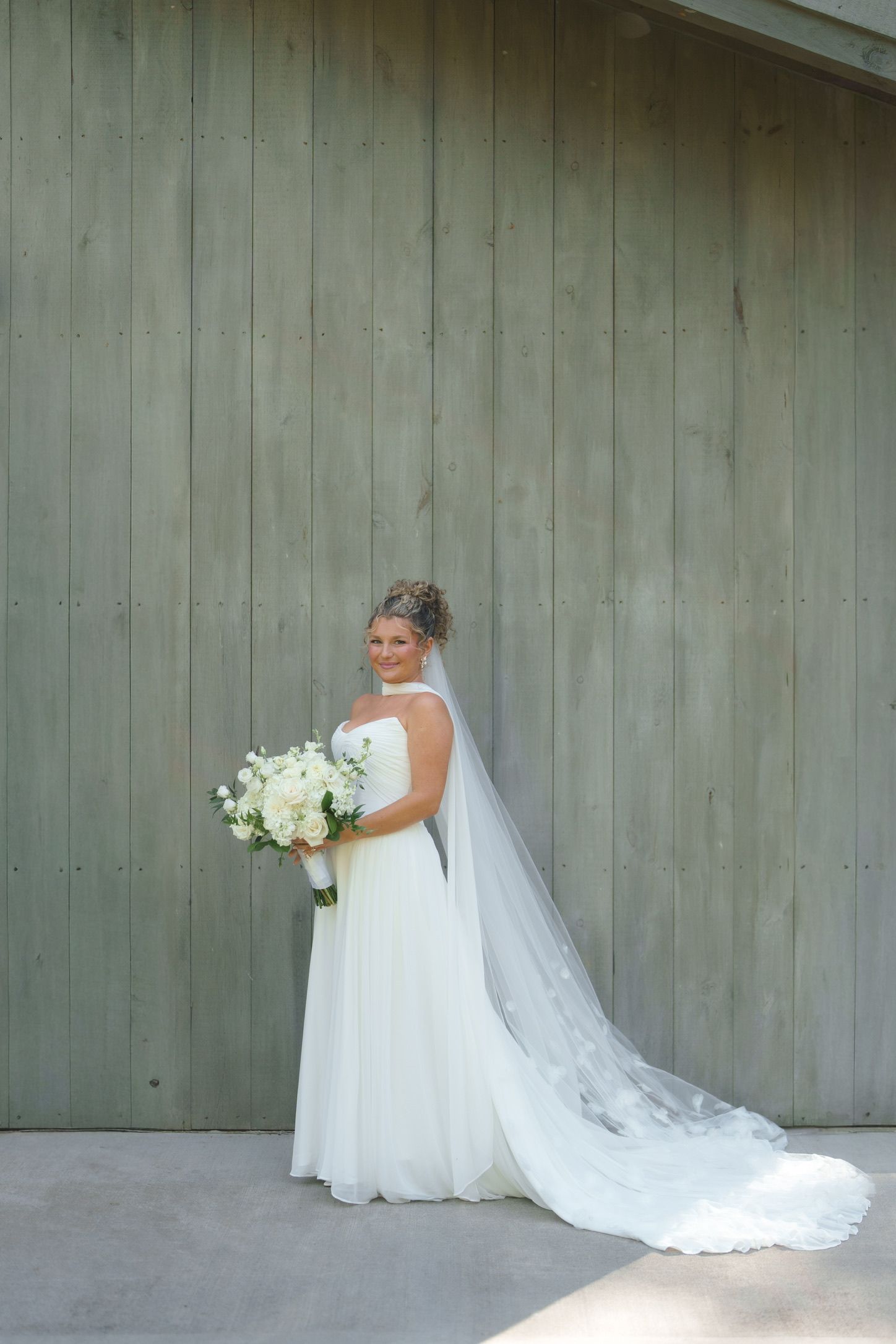 Bride in white gown and long veil holds white bouquet, posing in front of rustic green wooden barn doors.