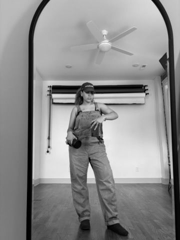 Black and white mirror selfie of a person in overalls holding a camera, with a ceiling fan visible in the background.