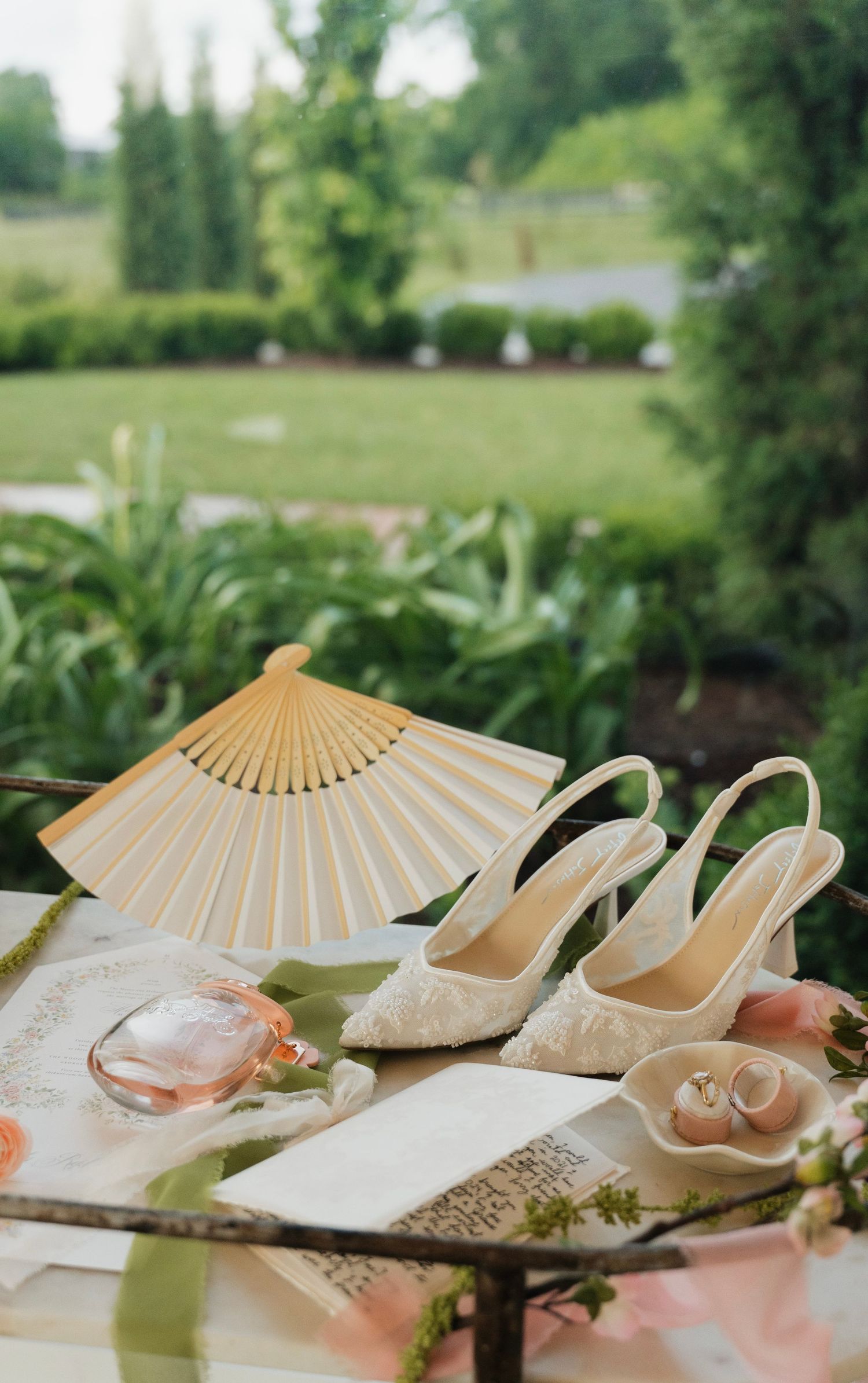 Elegant bridal flat lay with lace heels, gold fan, perfume, rings, and florals on a table with lush garden backdrop.