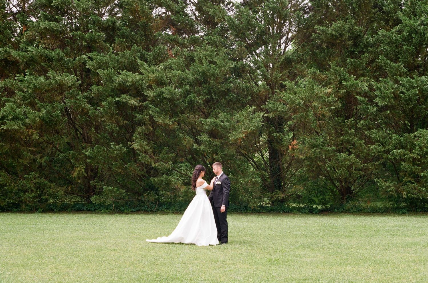 A bride and groom share a romantic moment on a lush green lawn surrounded by tall evergreen trees.