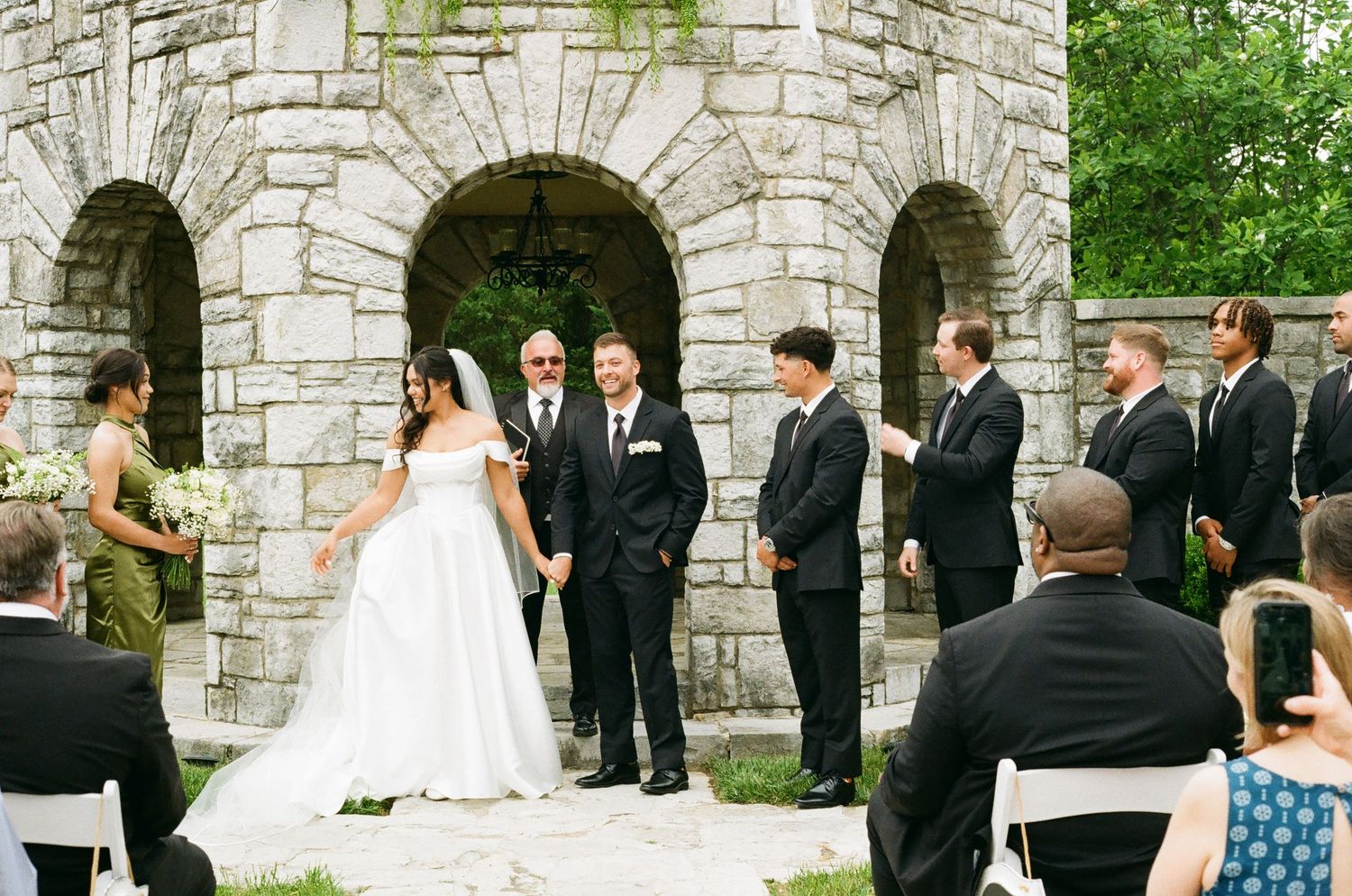 Outdoor wedding ceremony at a stone castle venue with bride in white gown, groom, and groomsmen in black suits.
