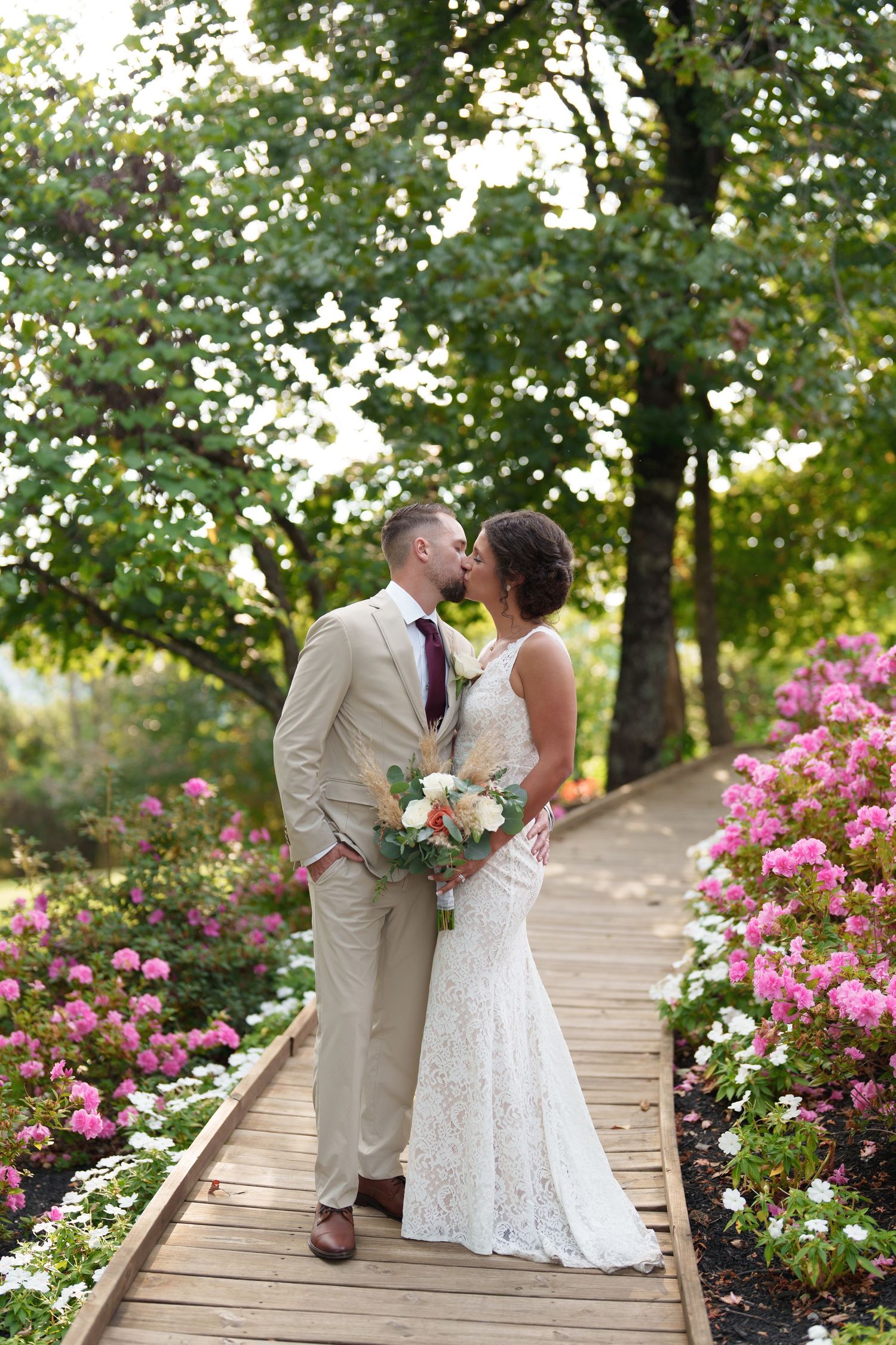Bride and groom kissing on a wooden boardwalk surrounded by pink roses and lush green trees in a garden.