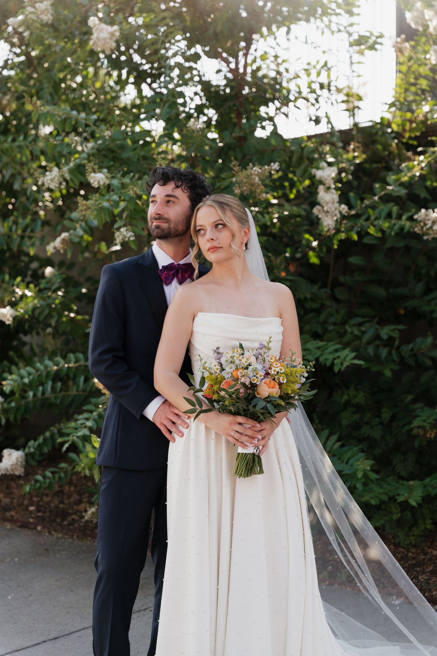 Bride in strapless white gown holding wildflower bouquet stands with groom in navy suit and burgundy bow tie in garden.