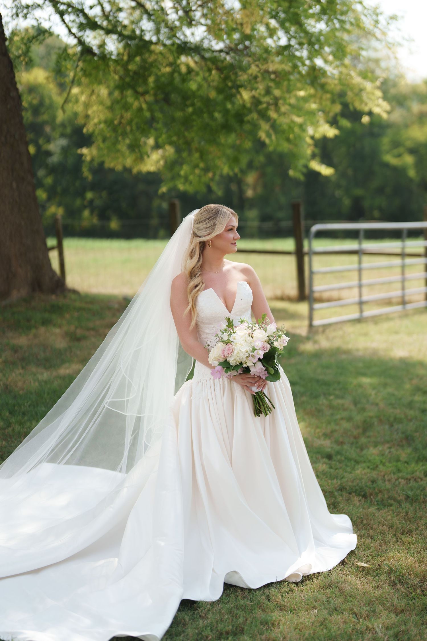 Bride in strapless white ball gown with long veil holds pink bouquet outdoors near wooden fence and green trees.