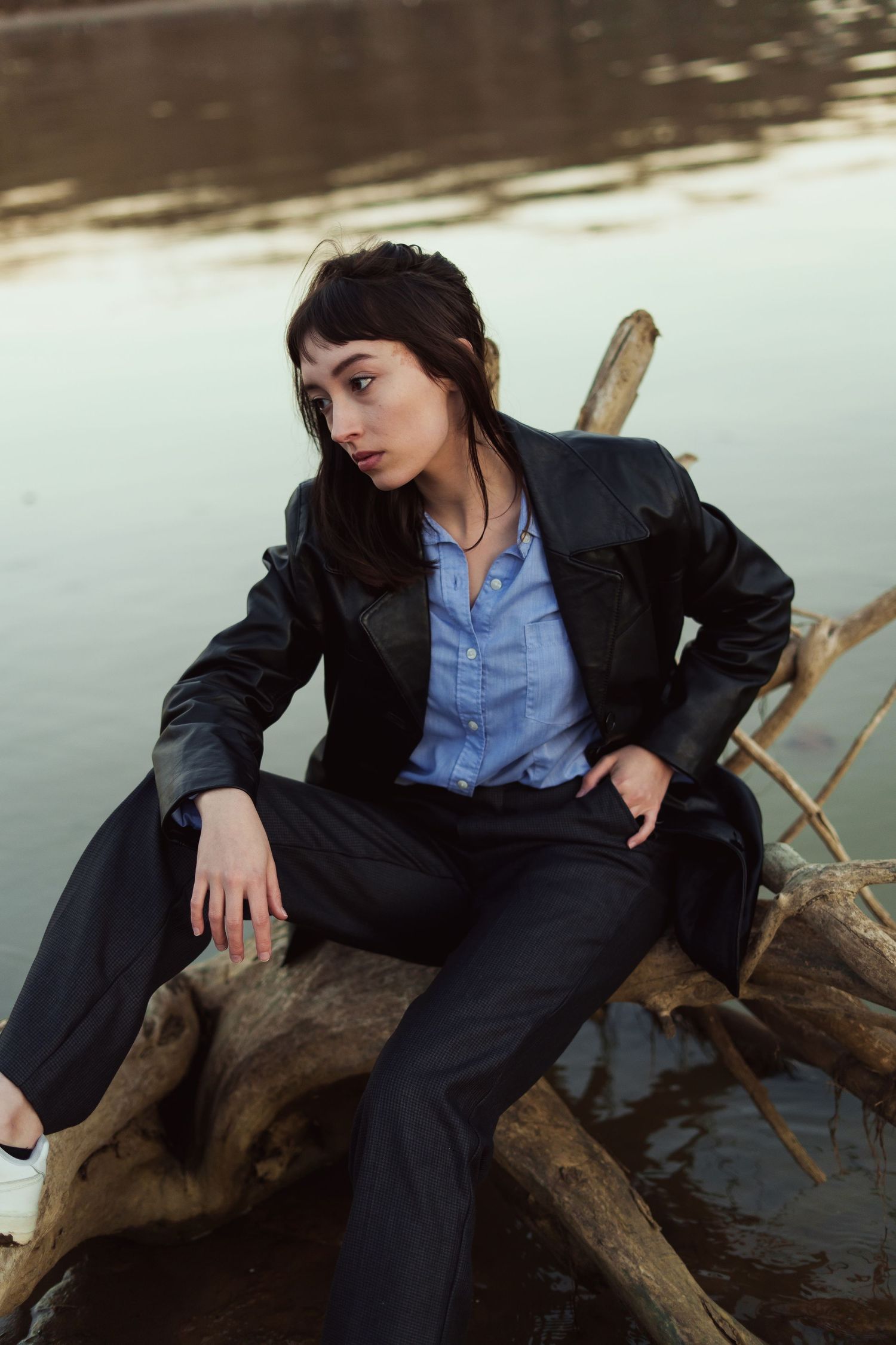 A person in business attire sits casually on driftwood by a tranquil lake during golden hour.