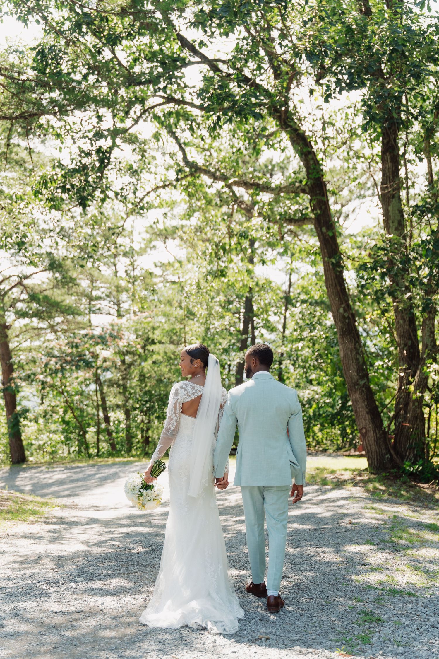 Bride and groom walking hand in hand down a gravel path surrounded by lush green trees on their wedding day.