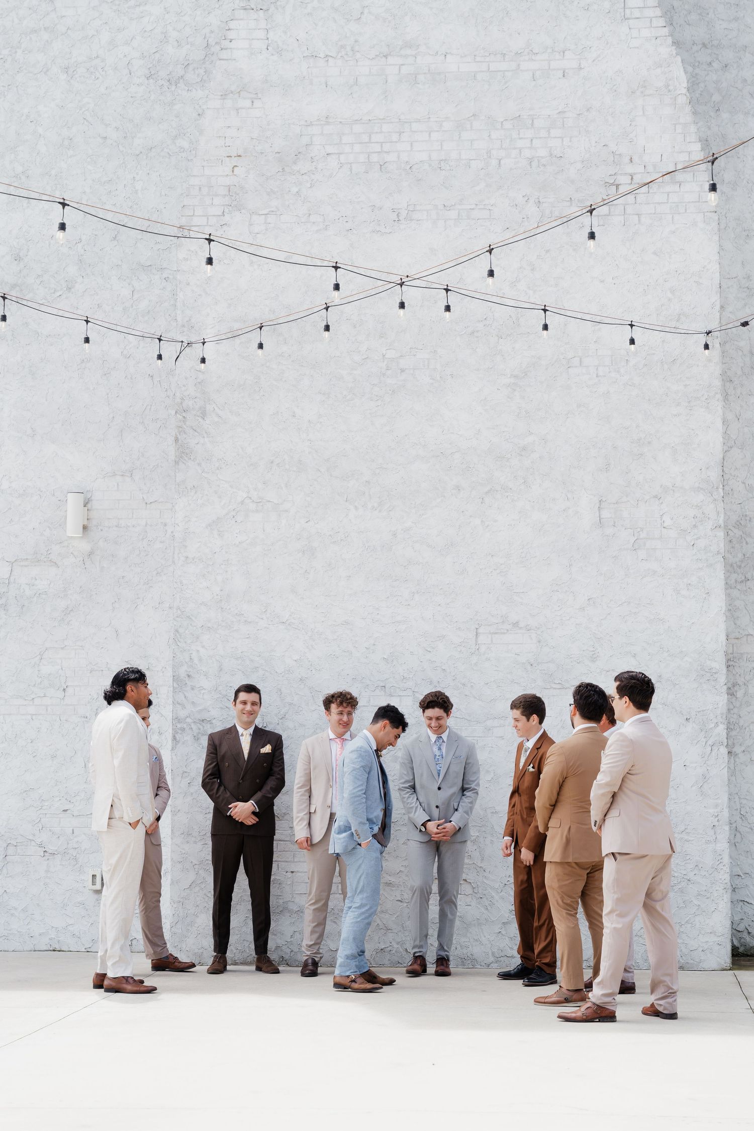 A group of groomsmen in mismatched neutral suits stand together against a white brick wall with string lights above.