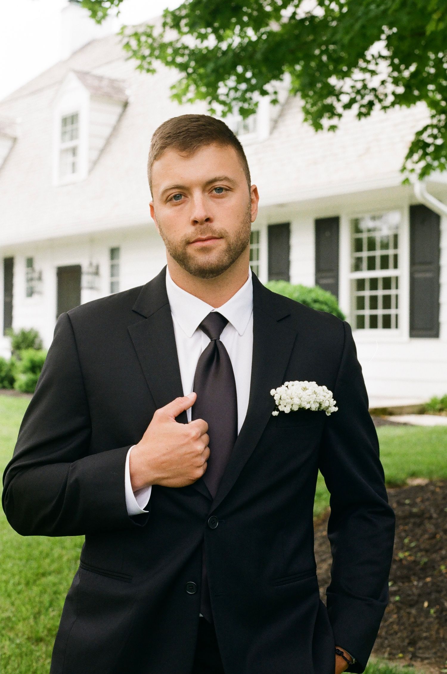 A groom in a black suit with white pocket square and dark tie stands confidently outside a white colonial home.
