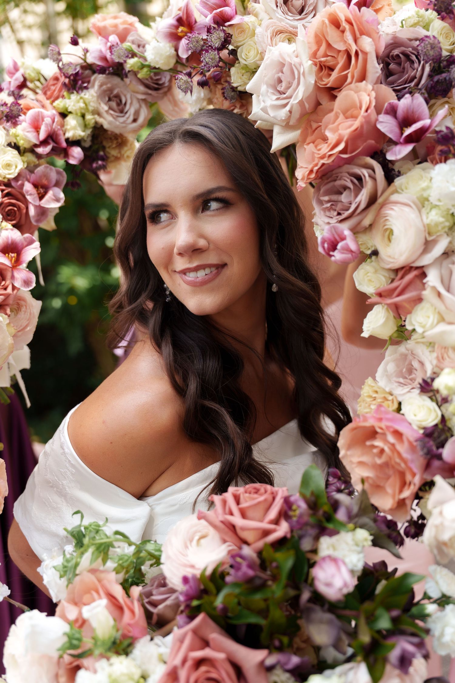 A bride poses framed by a stunning floral arch of pink, peach and white roses in an elegant outdoor setting.