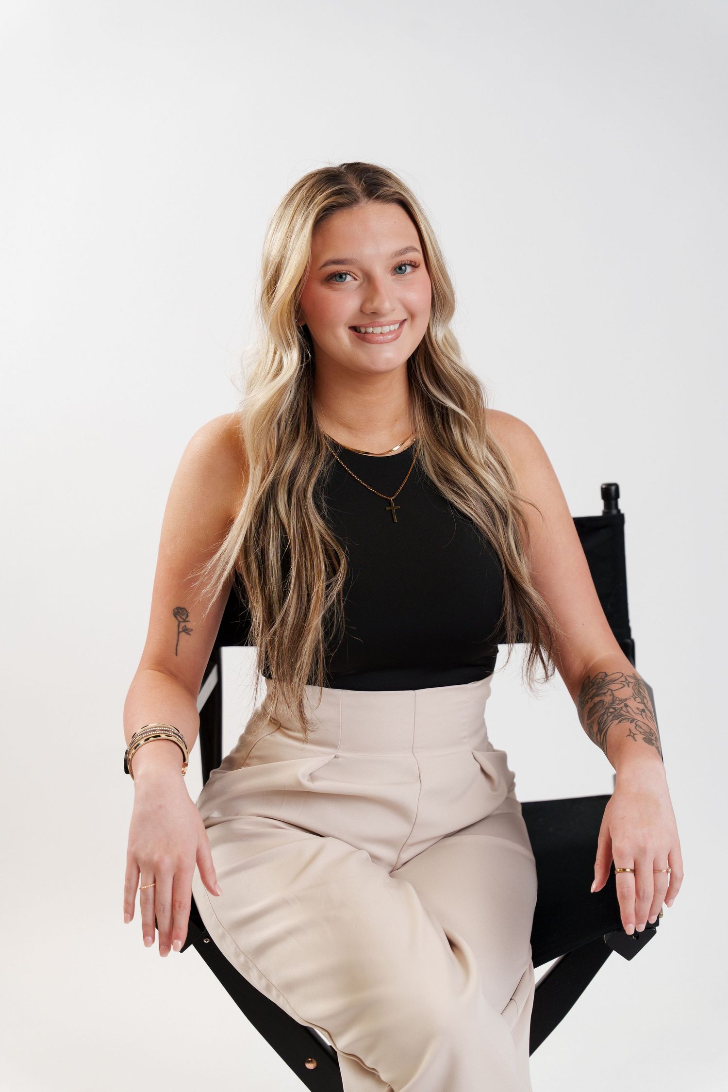 Professional studio portrait of someone in a black sleeveless top and beige pants sitting on a director's chair.