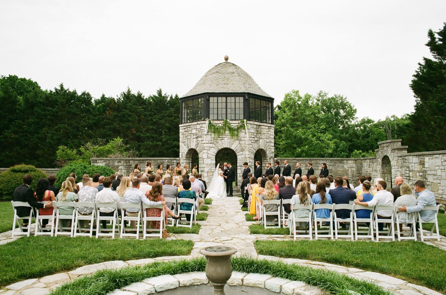 Outdoor wedding ceremony at a stone garden venue with white chairs and a decorative fountain in the foreground.