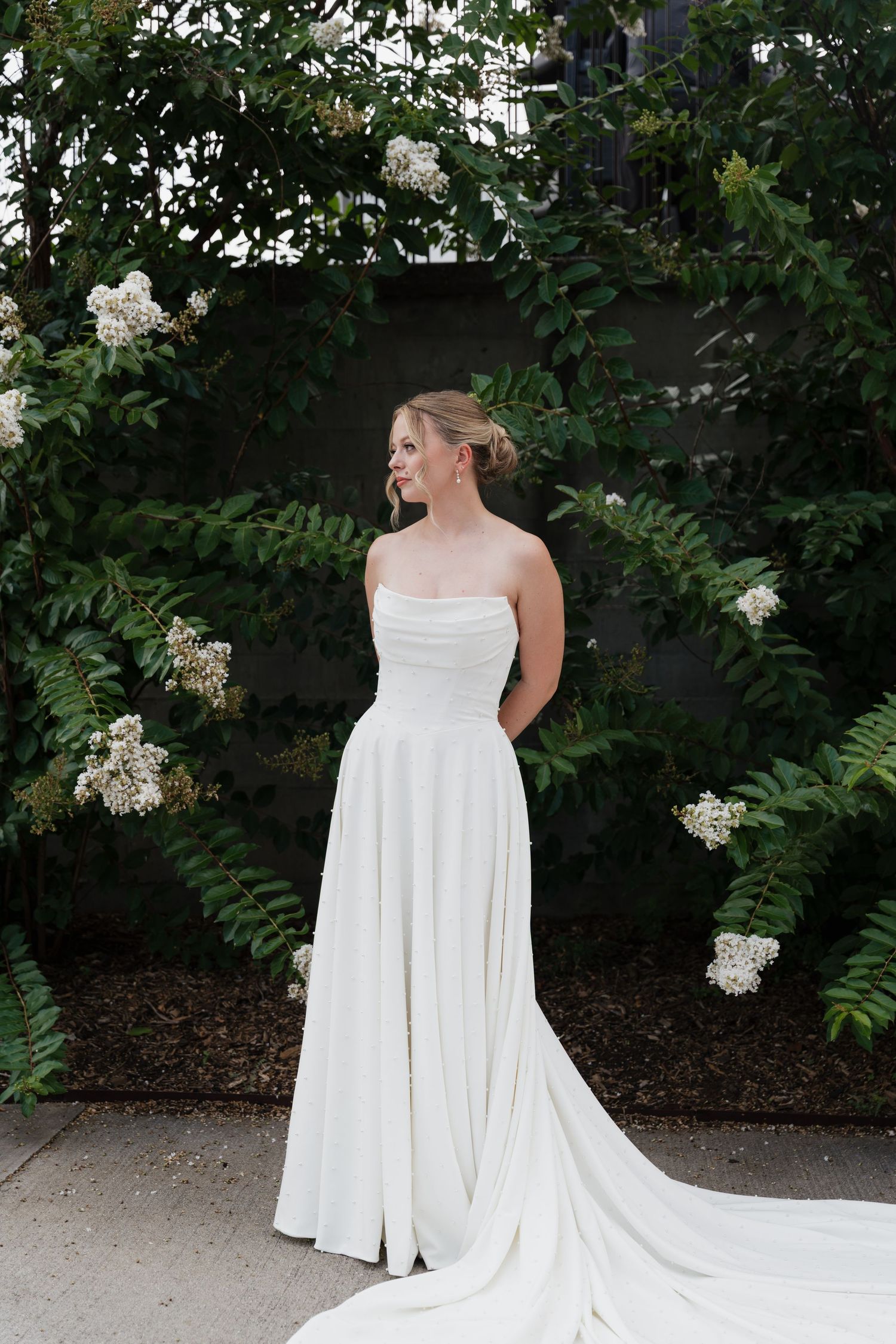 Bride in strapless white gown with train stands before lush green foliage and white blooms in elegant outdoor setting.