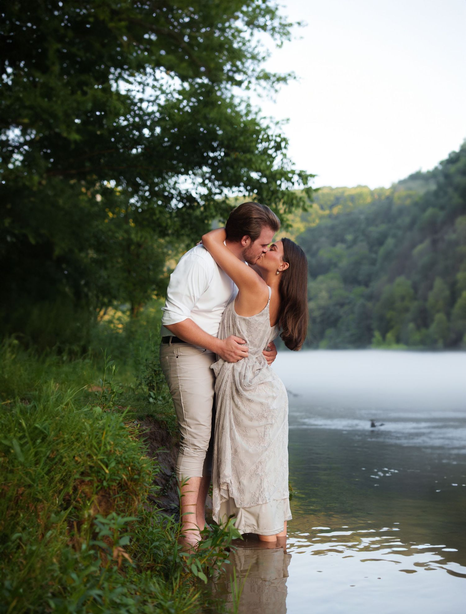 Couple sharing a romantic kiss while standing in a shallow river surrounded by lush green trees and misty hills.