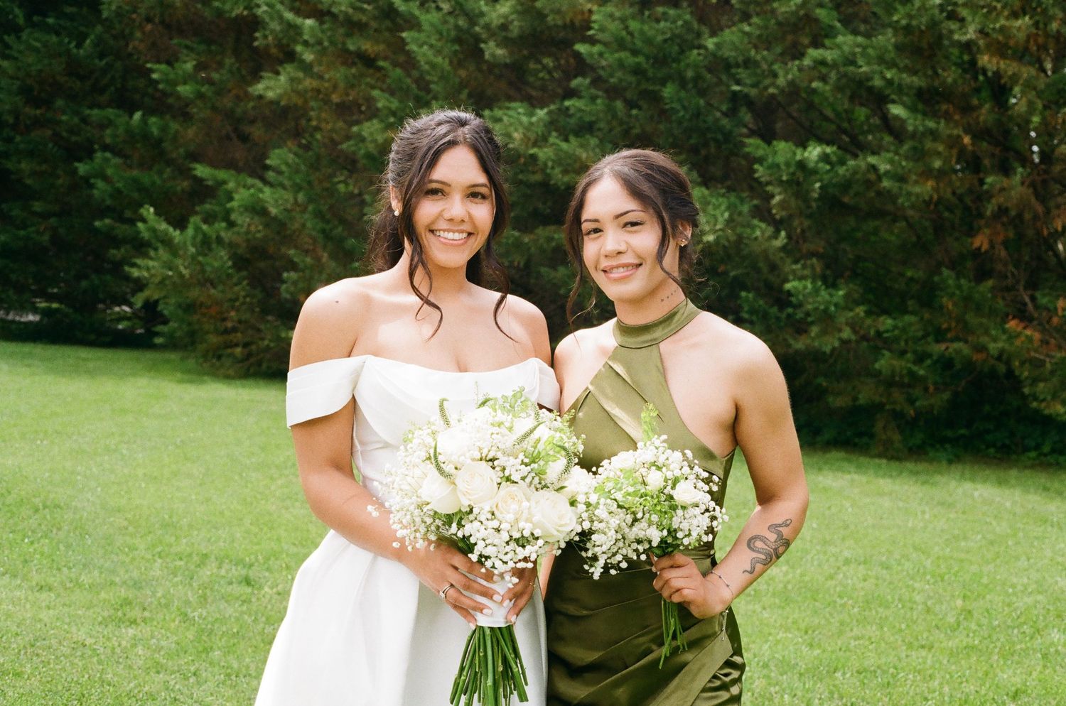 Bride in white off-shoulder gown and bridesmaid in olive green dress holding white bouquets outdoors.