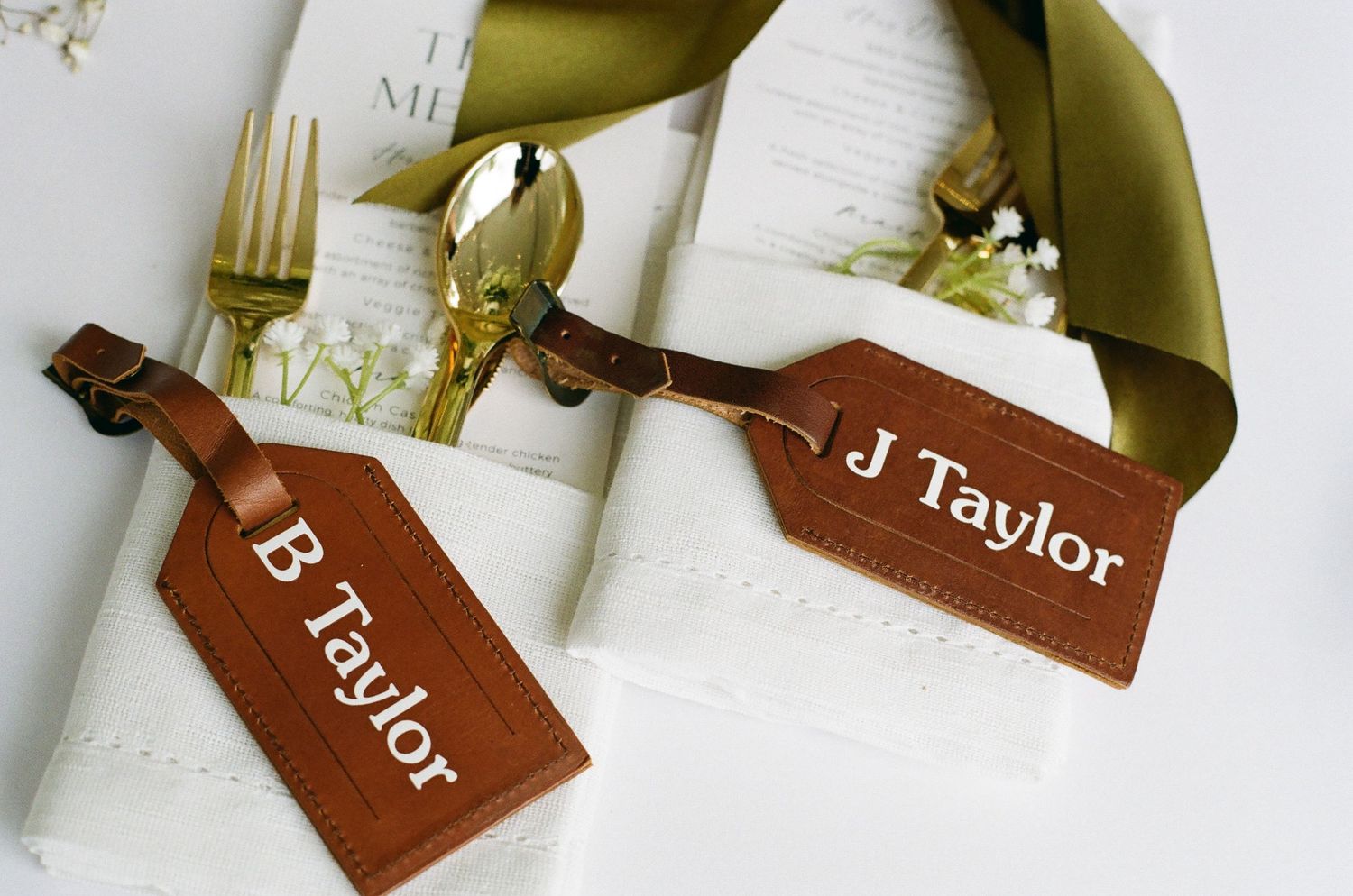 Personalized leather luggage tags with names B Taylor and J Taylor displayed on white napkins with gold ribbon and cutlery.