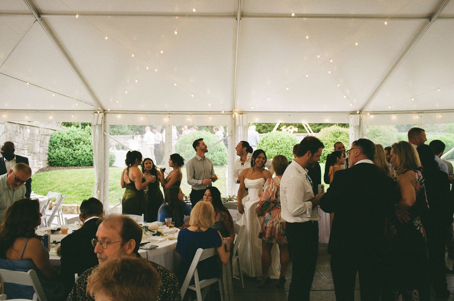 Elegant outdoor wedding reception under a white tent with string lights, guests mingling in formal attire.