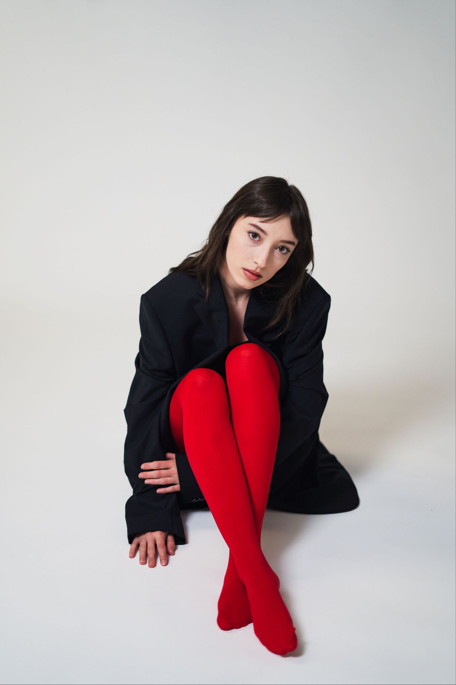 Figure in black jacket poses artistically against white backdrop with vibrant red stockings creating dramatic contrast.