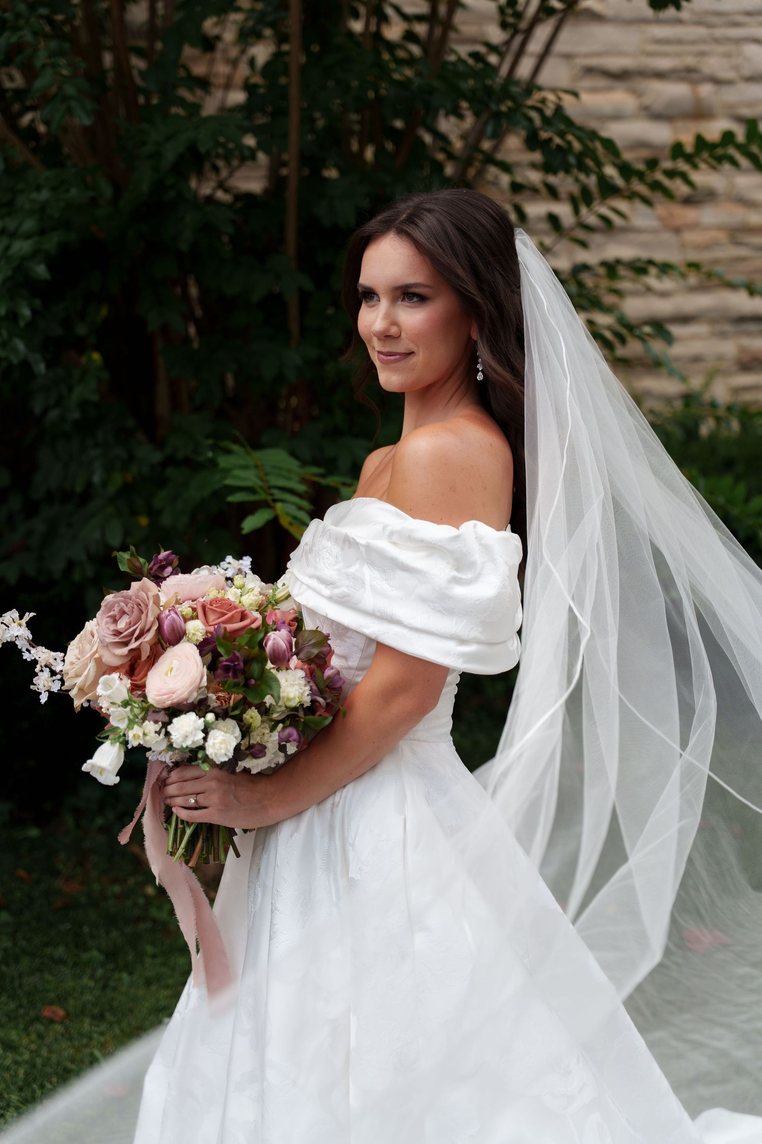 A bride in a white gown with flowing veil poses gracefully against a natural outdoor backdrop.