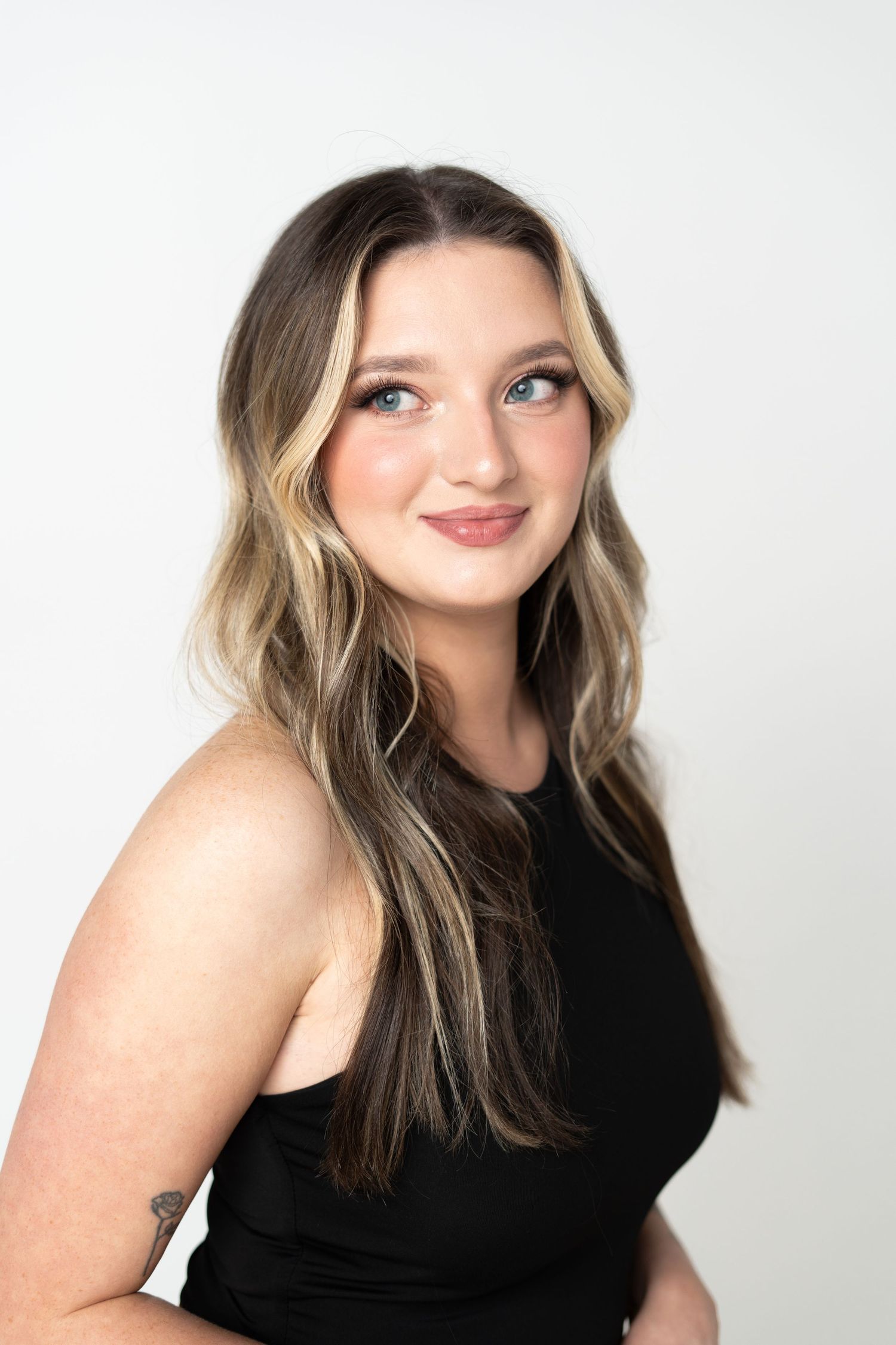 Professional portrait of someone with long wavy blonde highlights wearing a black sleeveless top against white background.