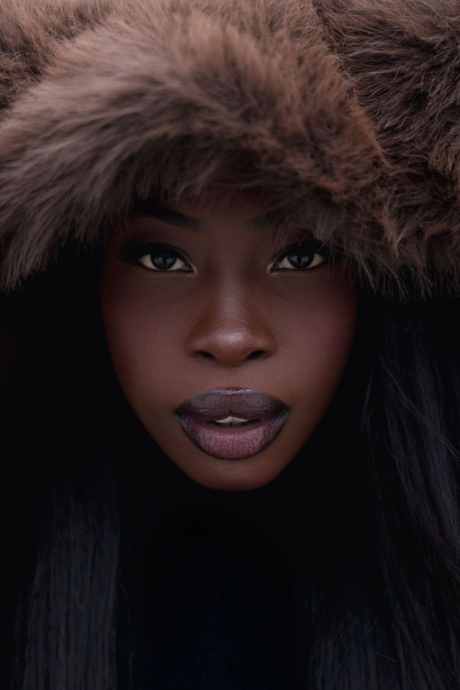 A striking Black woman with dark lipstick and blue eyes wearing a brown fur hood against a dark background.