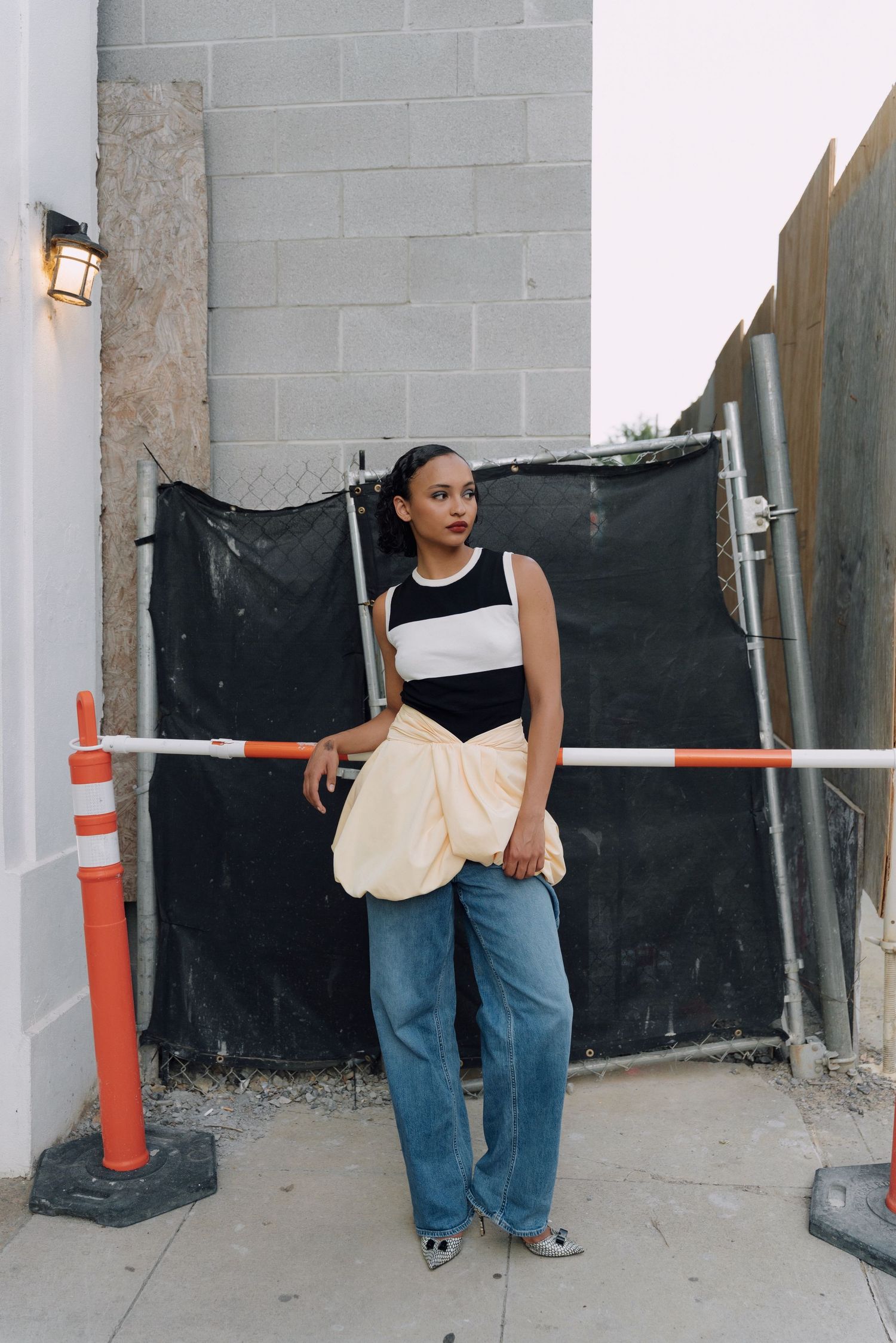 Woman in black and white top, cream bag, and wide-leg jeans poses on sidewalk near construction barrier.