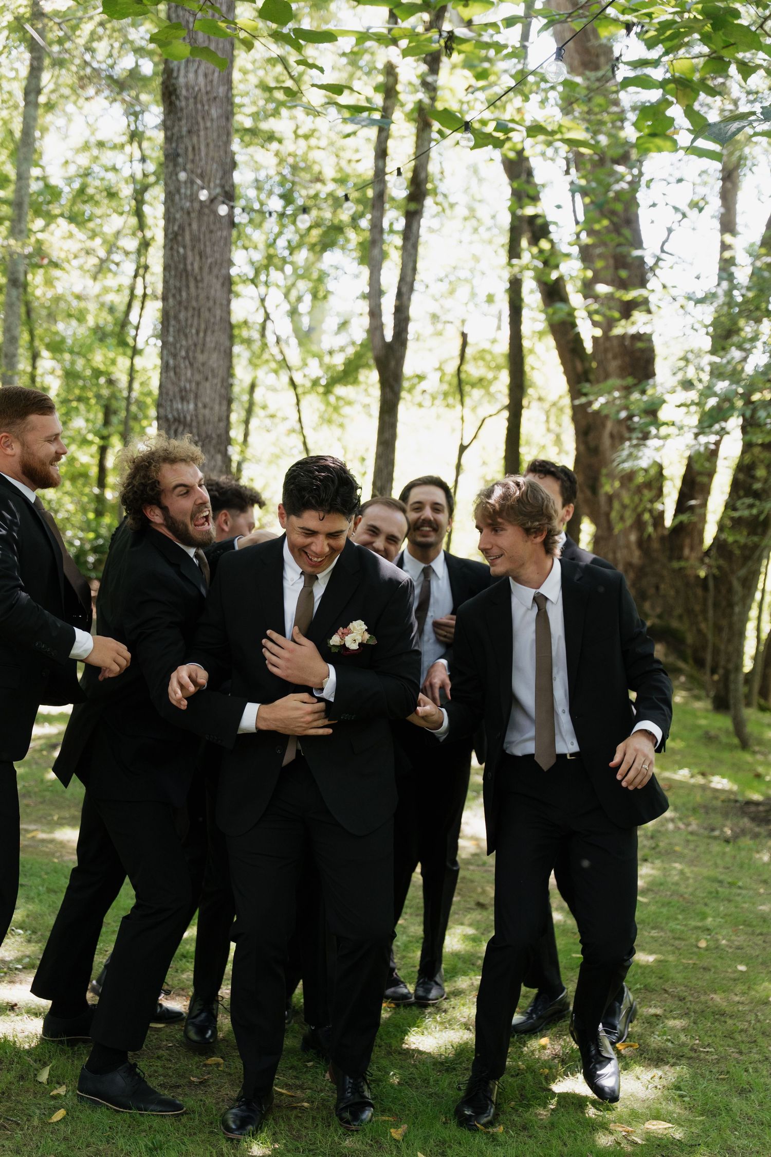 Groomsmen in black suits laughing and celebrating outdoors in a wooded setting during a wedding.