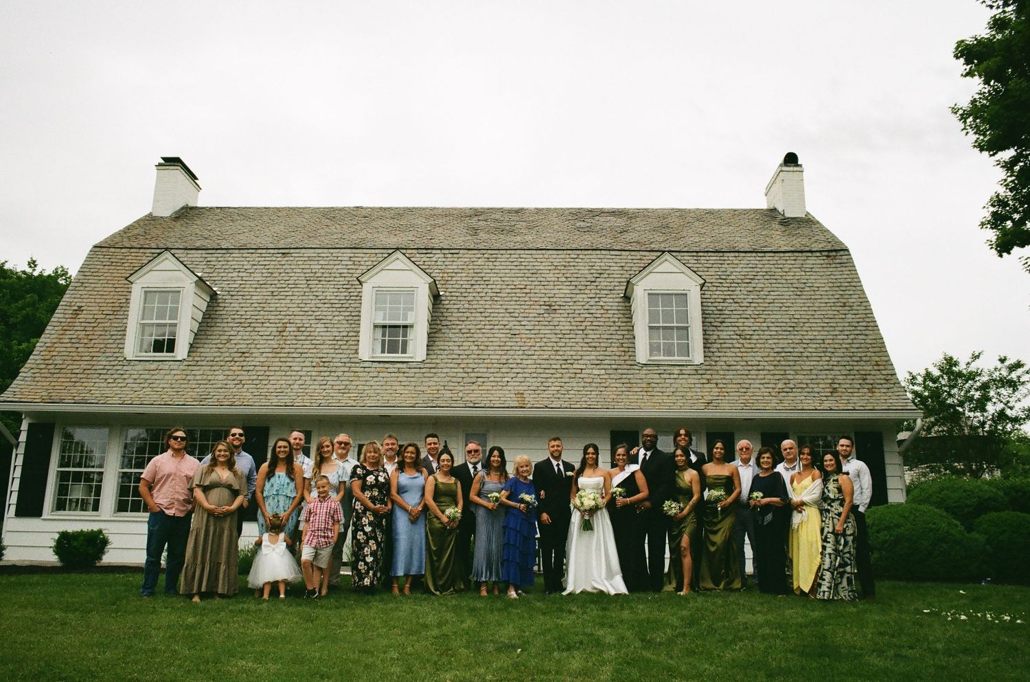 Wedding party poses in front of a charming cottage-style venue with shingled roof on a cloudy day.