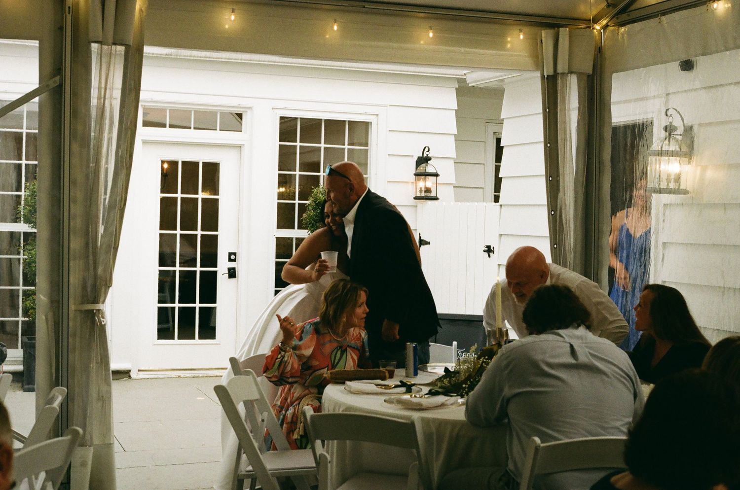 Wedding reception guests seated at tables on white decorated patio at night with string lights overhead.