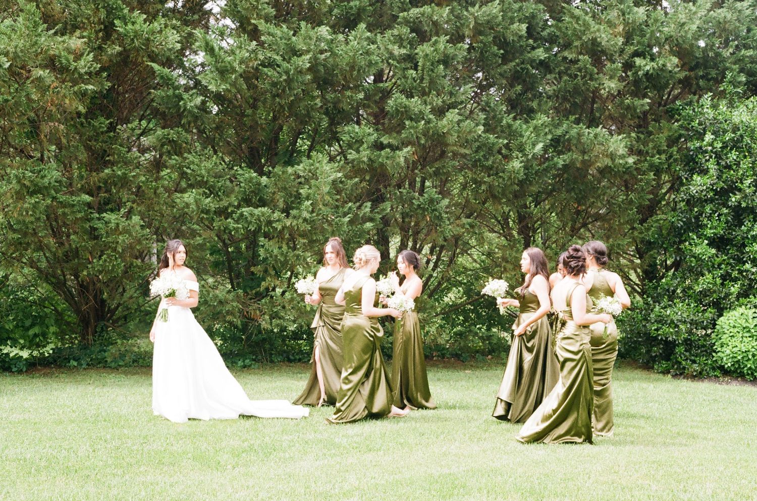 Bride in white gown with bridesmaids in olive green dresses holding bouquets on lush green lawn with trees.