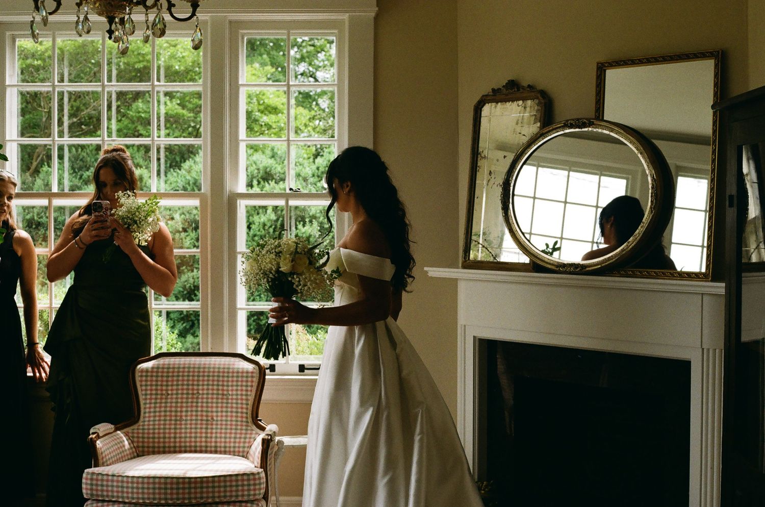 Bride in white off-shoulder gown holds bouquet near fireplace as bridesmaid photographs her in elegant room.