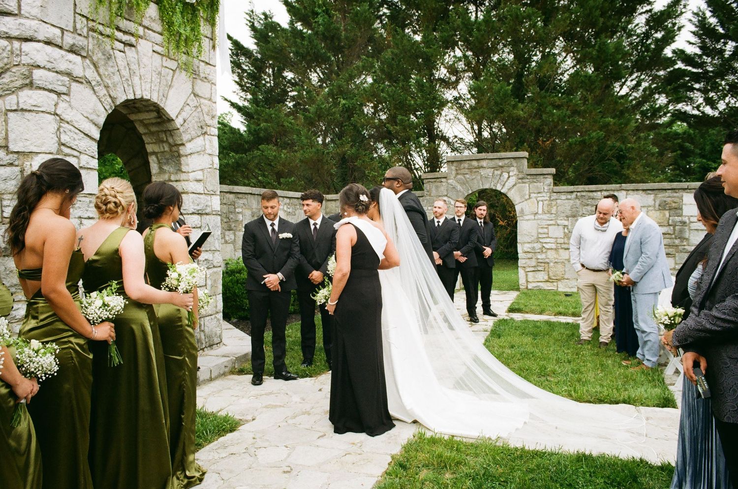 Outdoor wedding ceremony at a stone chapel with bridesmaids in olive green gowns and groomsmen in black suits.