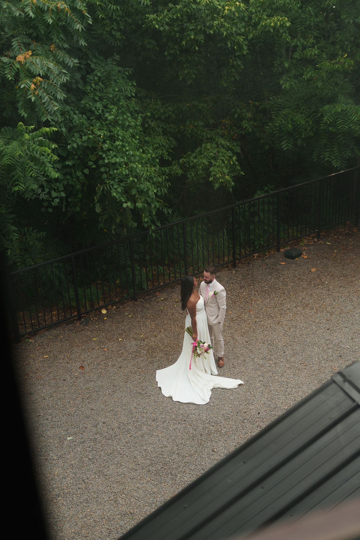 Aerial view of bride in white gown and groom in tan suit sharing a romantic moment on gravel path surrounded by lush greenery.
