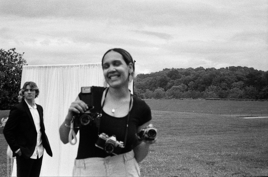 Woman smiling outdoors holding cameras at an event, black and white photo with trees and white draping in background.