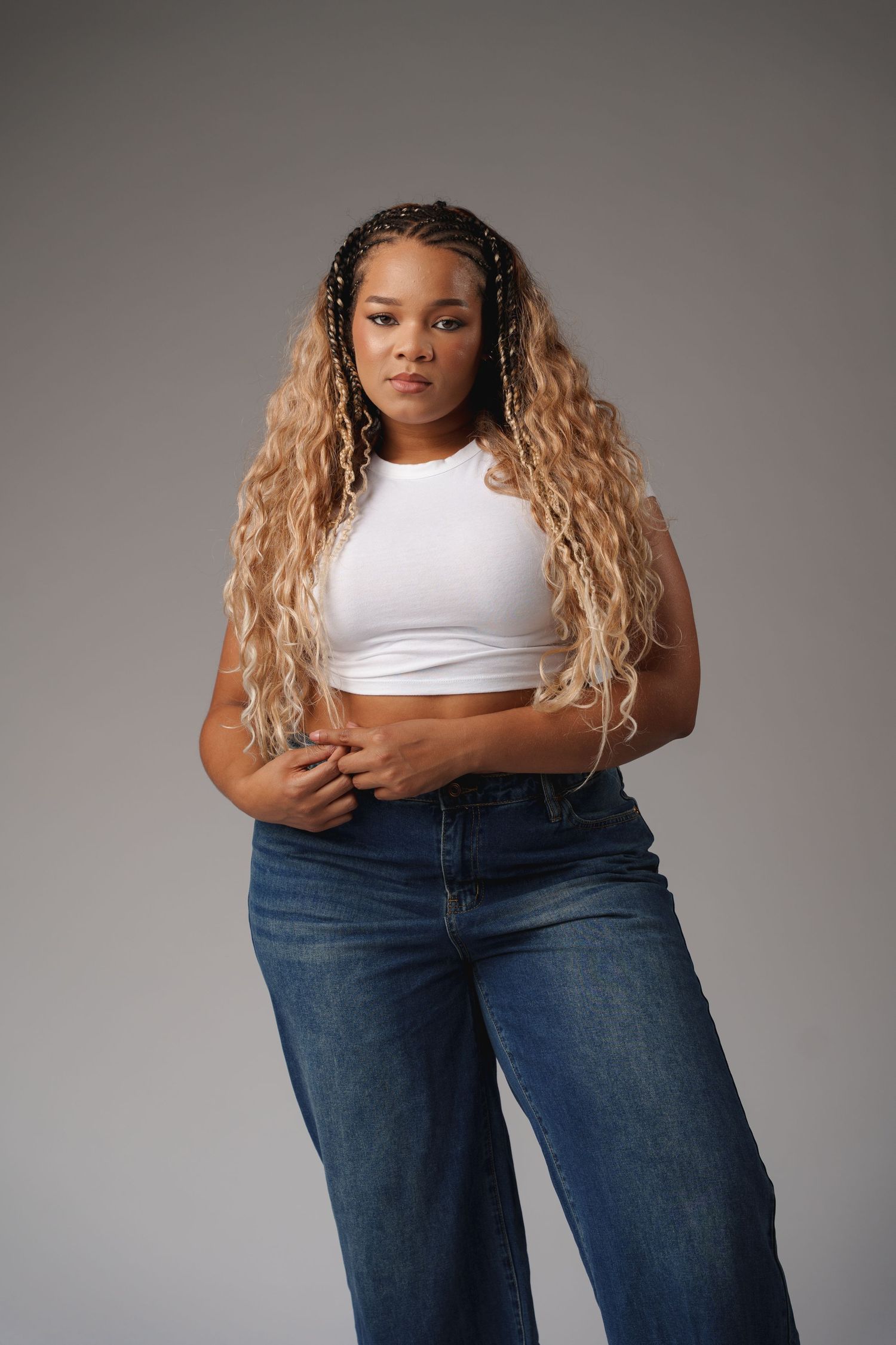 A person in a white crop top and relaxed-fit jeans poses confidently against a gray studio backdrop.