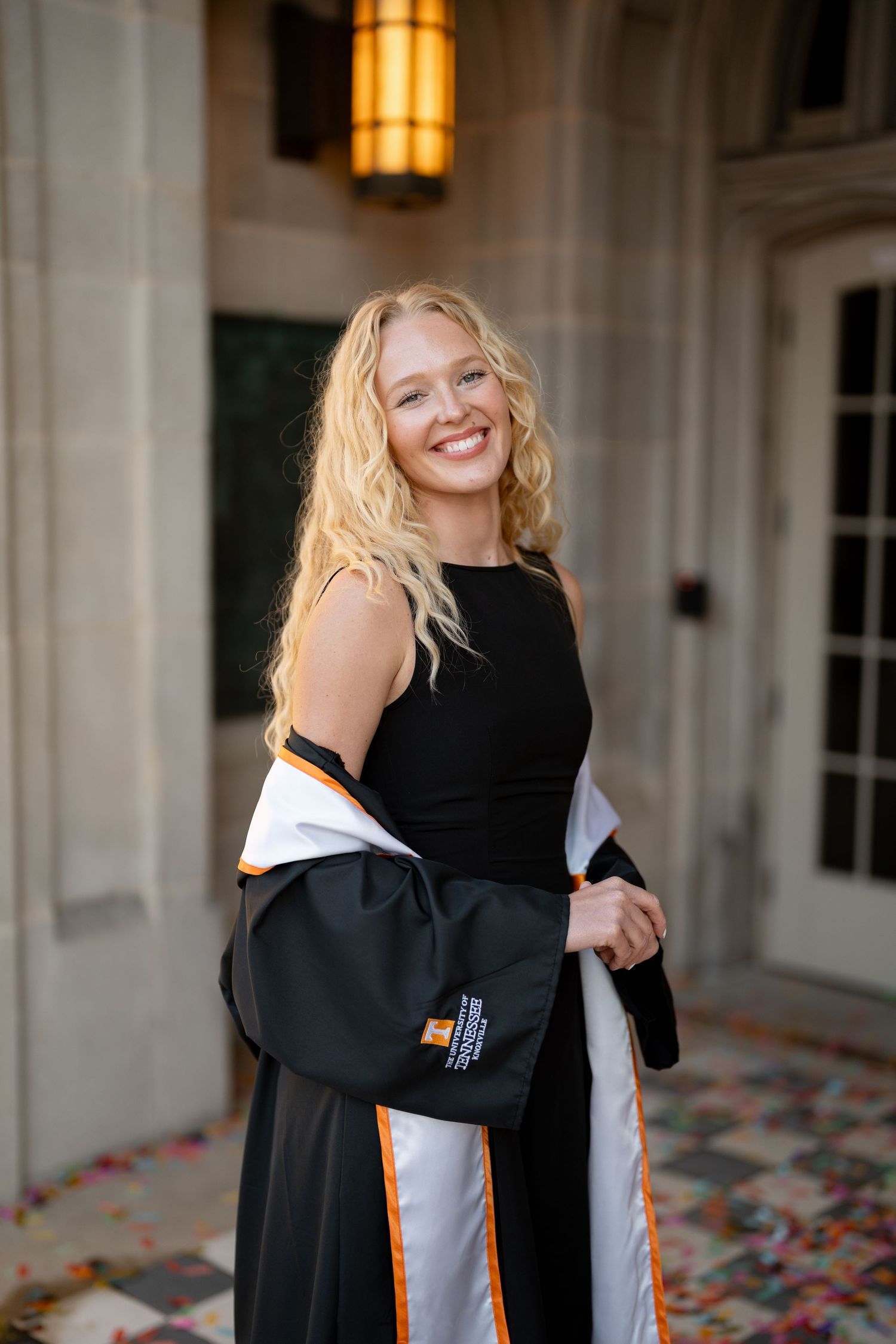 Smiling blonde woman in black graduation gown with orange and white trim poses in an elegant stone building.
