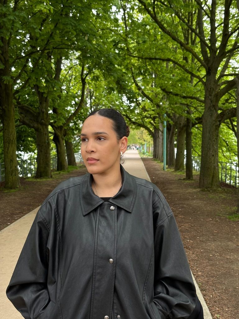 Woman in black leather jacket standing on tree-lined pathway in a park, looking thoughtfully to the side.