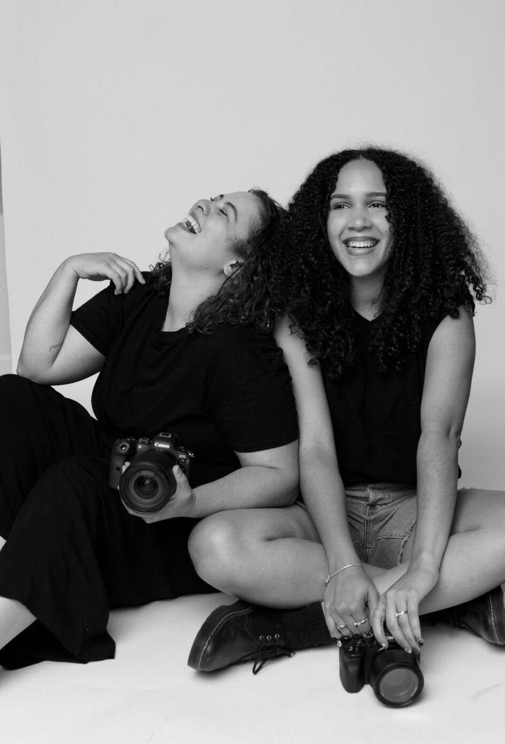 Two young women laughing and sitting on floor with cameras in black and white photo.