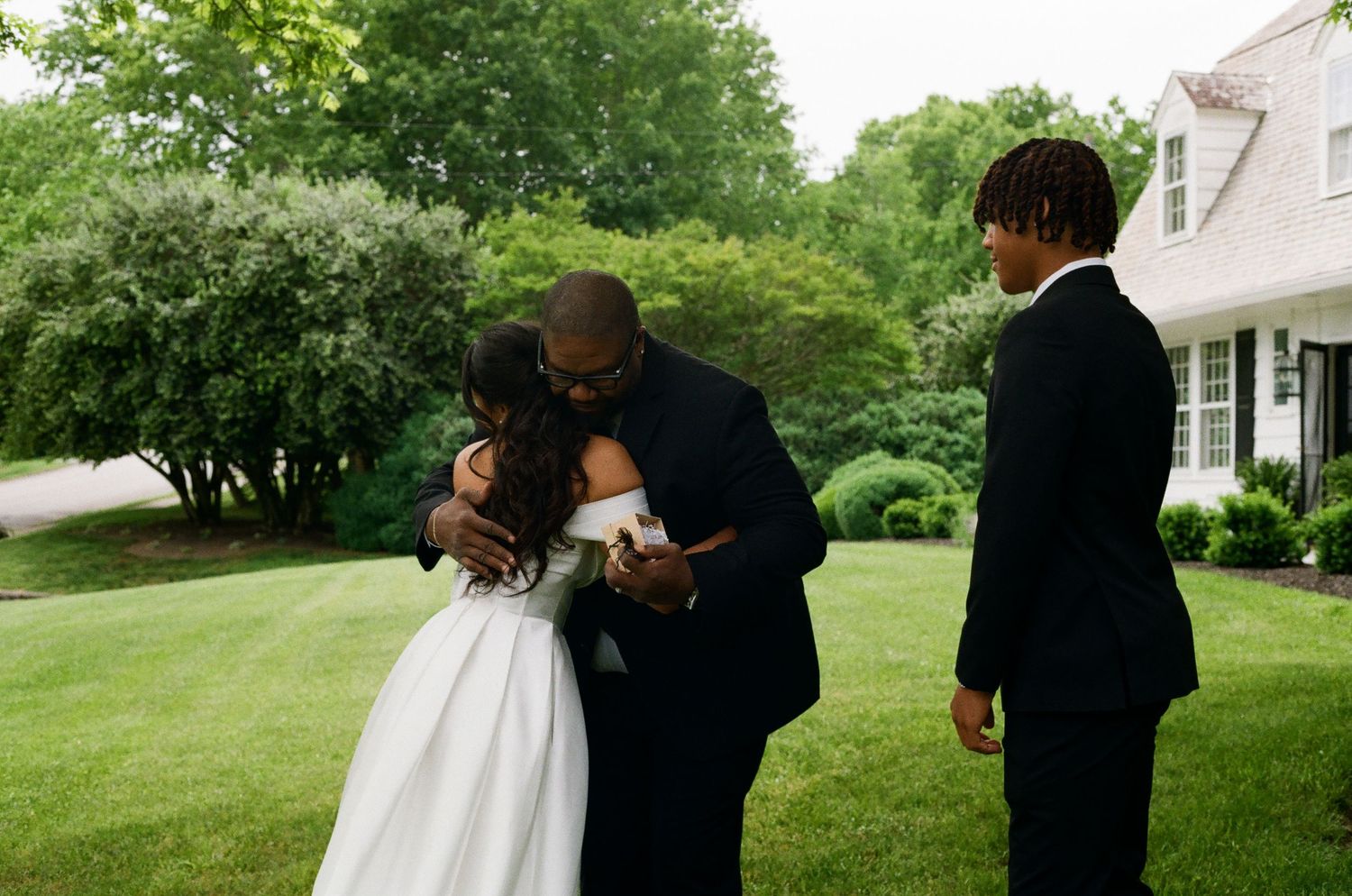 A groom embraces his bride in a white wedding dress outdoors on a green lawn while a young man in a suit stands nearby.