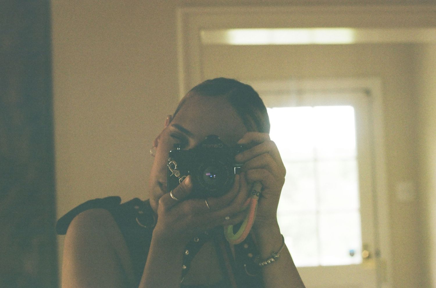 A woman takes a self-portrait in a mirror using a vintage film camera, wearing bracelets, in a warmly lit room.