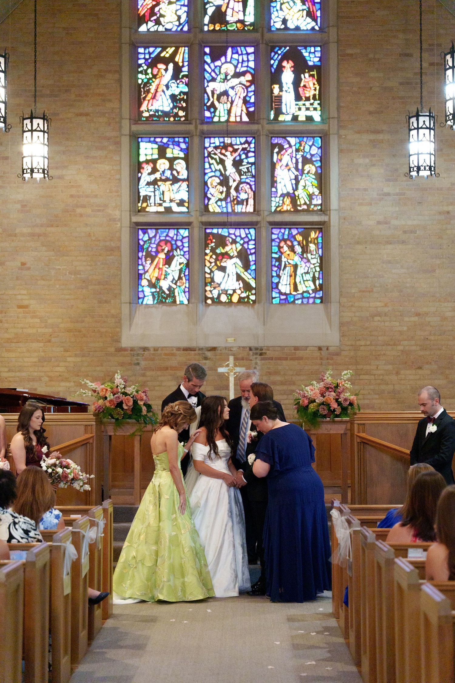 Church wedding ceremony with stained glass windows and wooden pews decorated with pink floral arrangements.