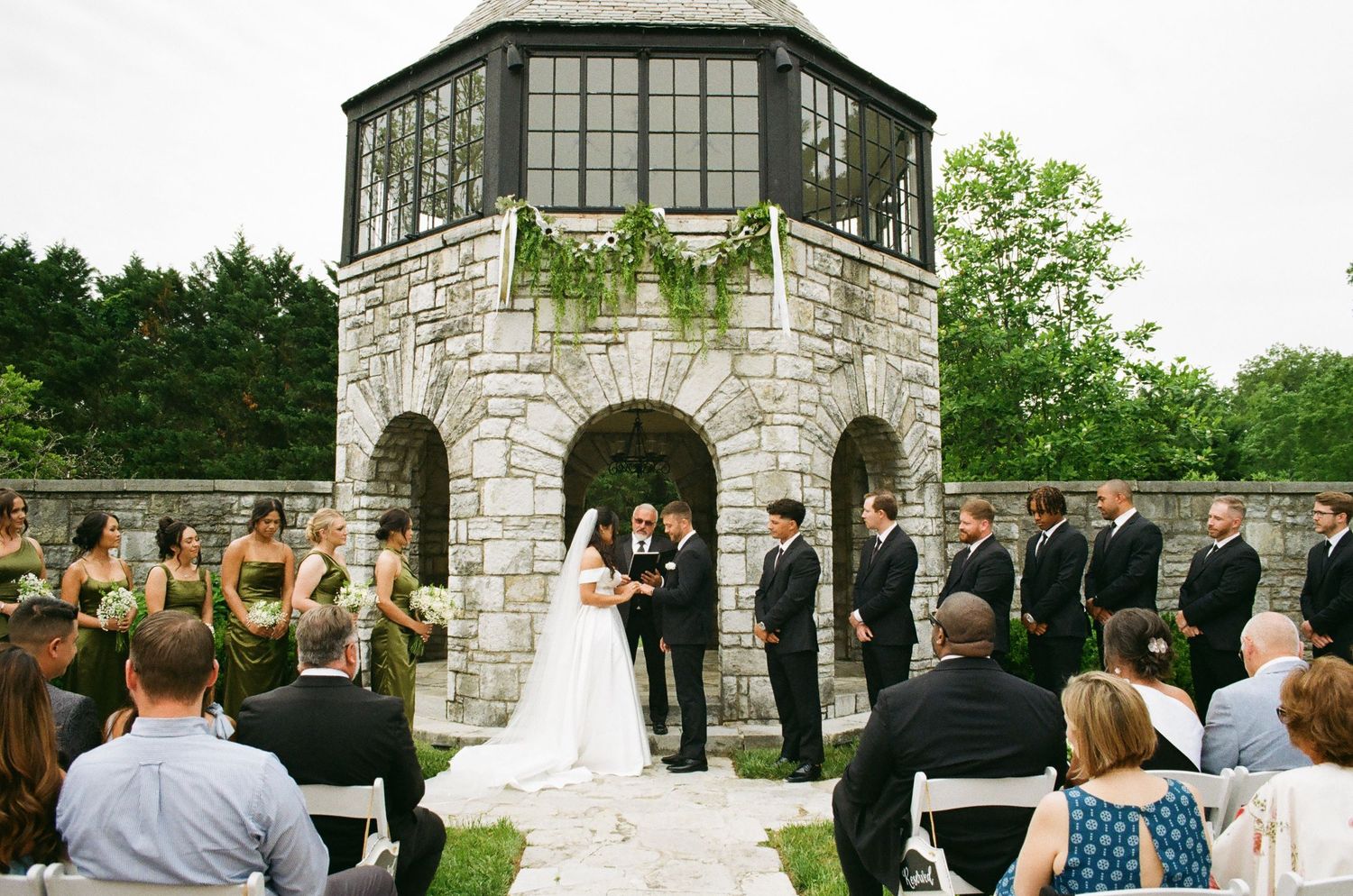 Outdoor wedding ceremony in front of a stone tower with bridal party and guests seated on white chairs.