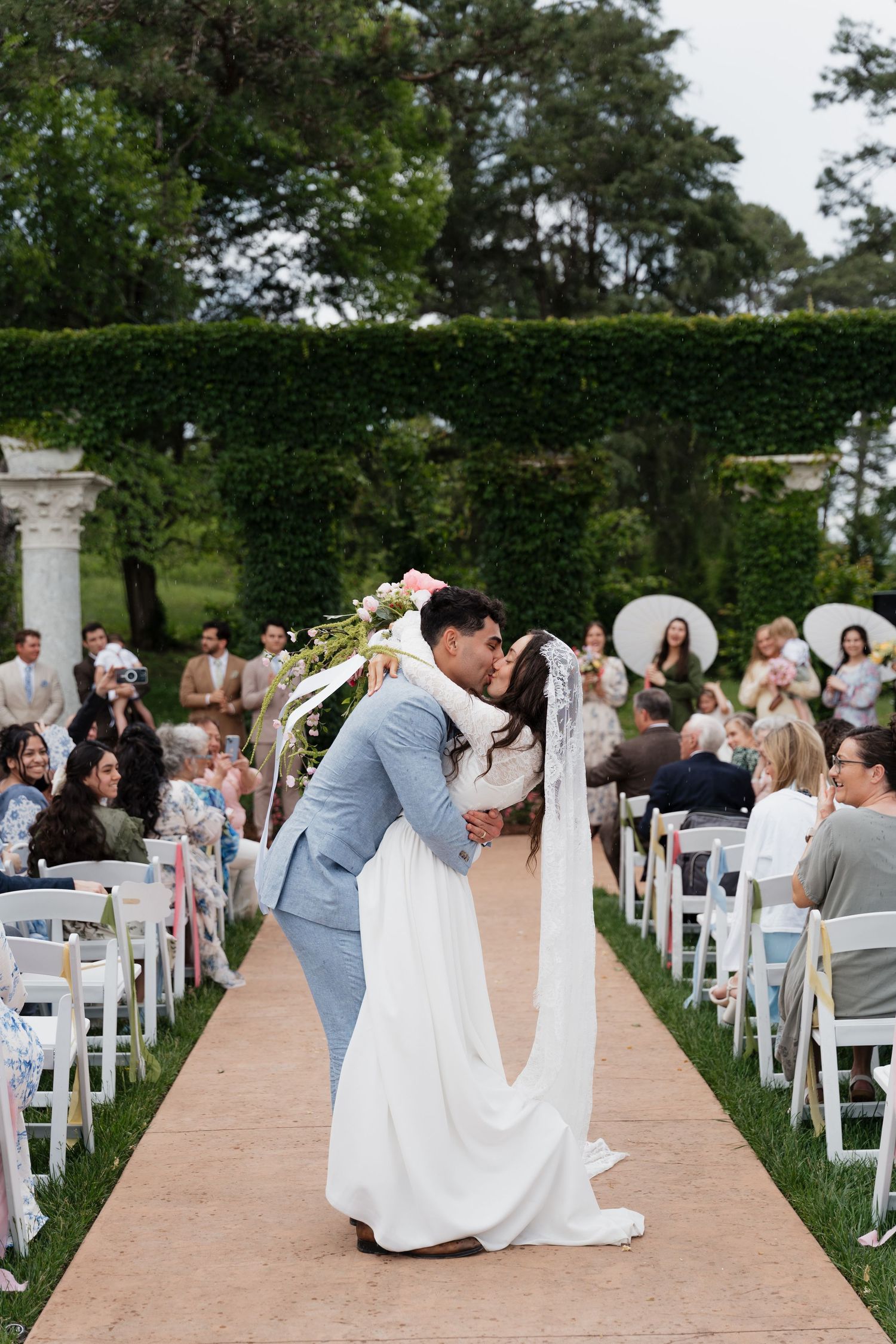 Bride and groom share first kiss at outdoor wedding ceremony aisle surrounded by guests and lush green hedges.
