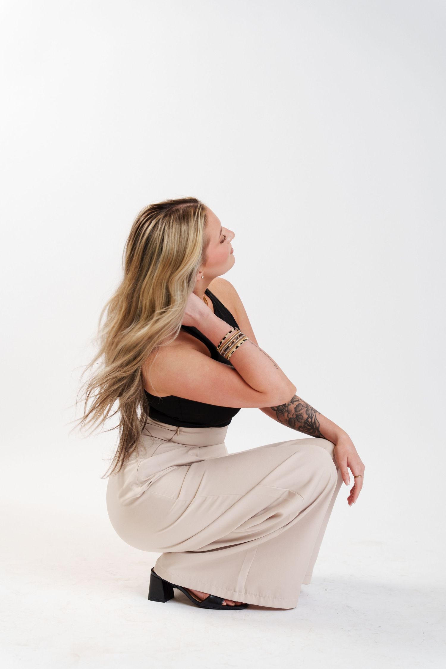 A woman in a black top and beige pants crouches down in a profile pose against a white backdrop.