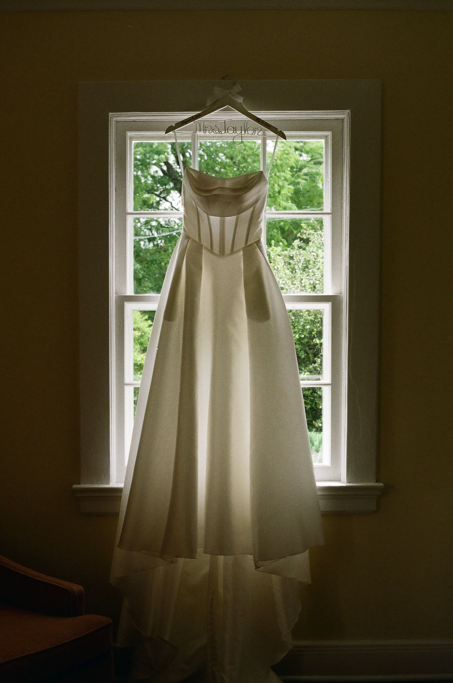 Elegant strapless white wedding gown hanging on a decorative hanger in a sunlit window with green trees outside.