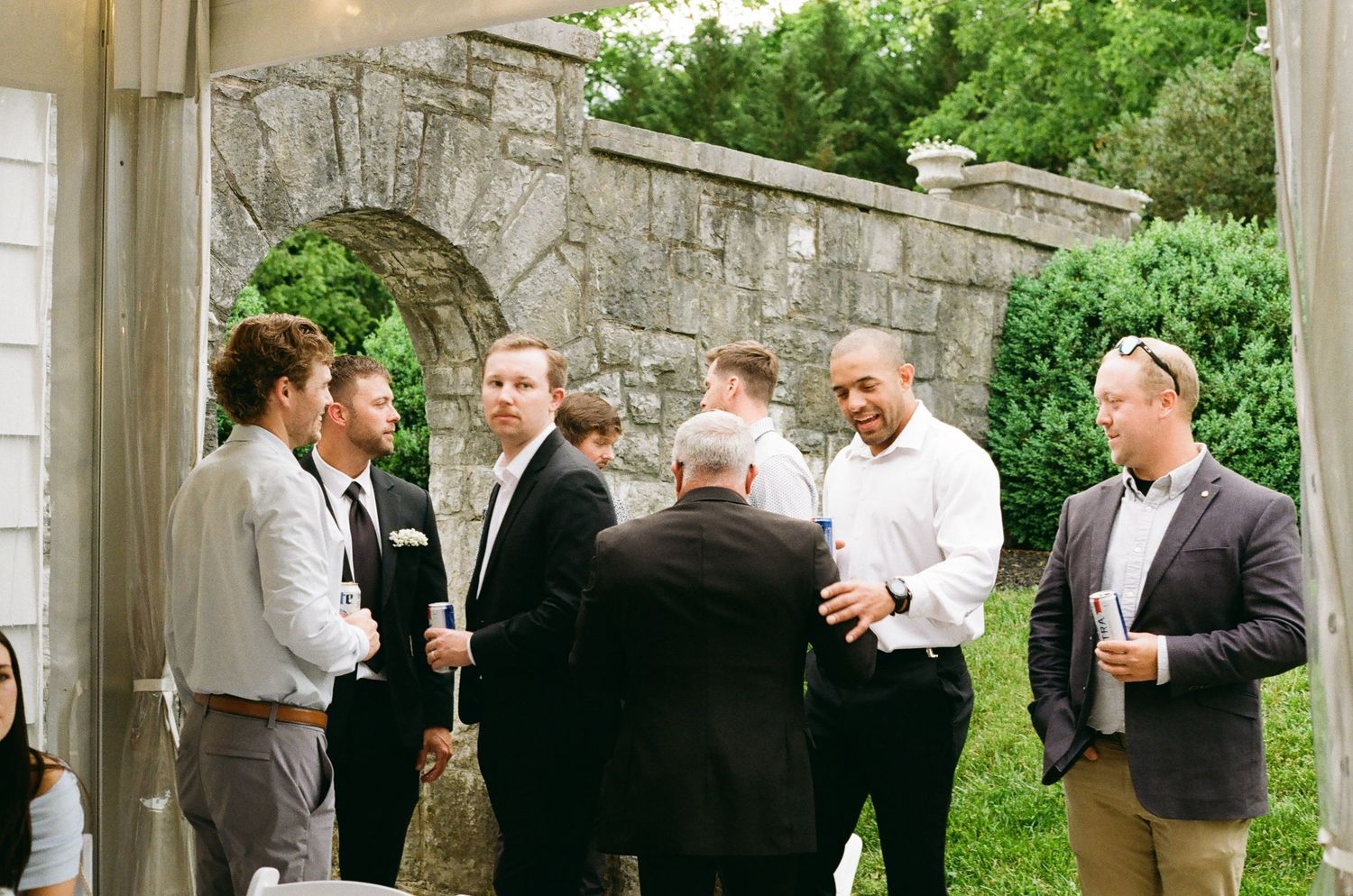 Group of men in semi-formal attire socializing outdoors near a stone arch wall at a wedding reception.