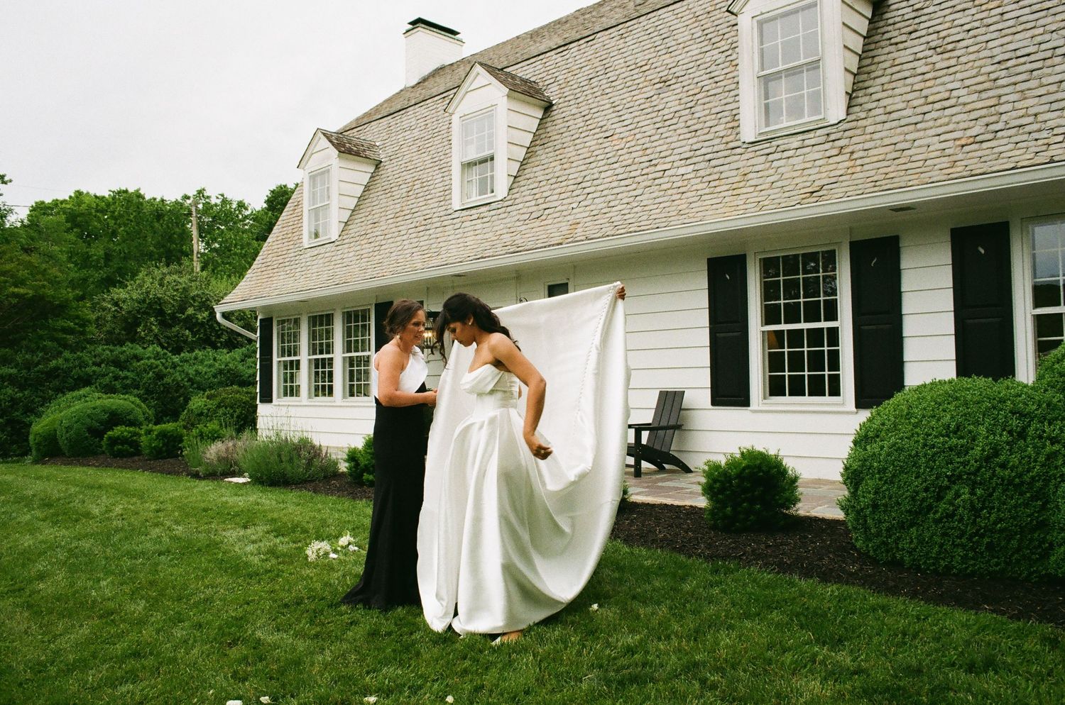 Bride in white gown with veil being adjusted by bridesmaid outside elegant white Cape Cod home with lush green lawn.