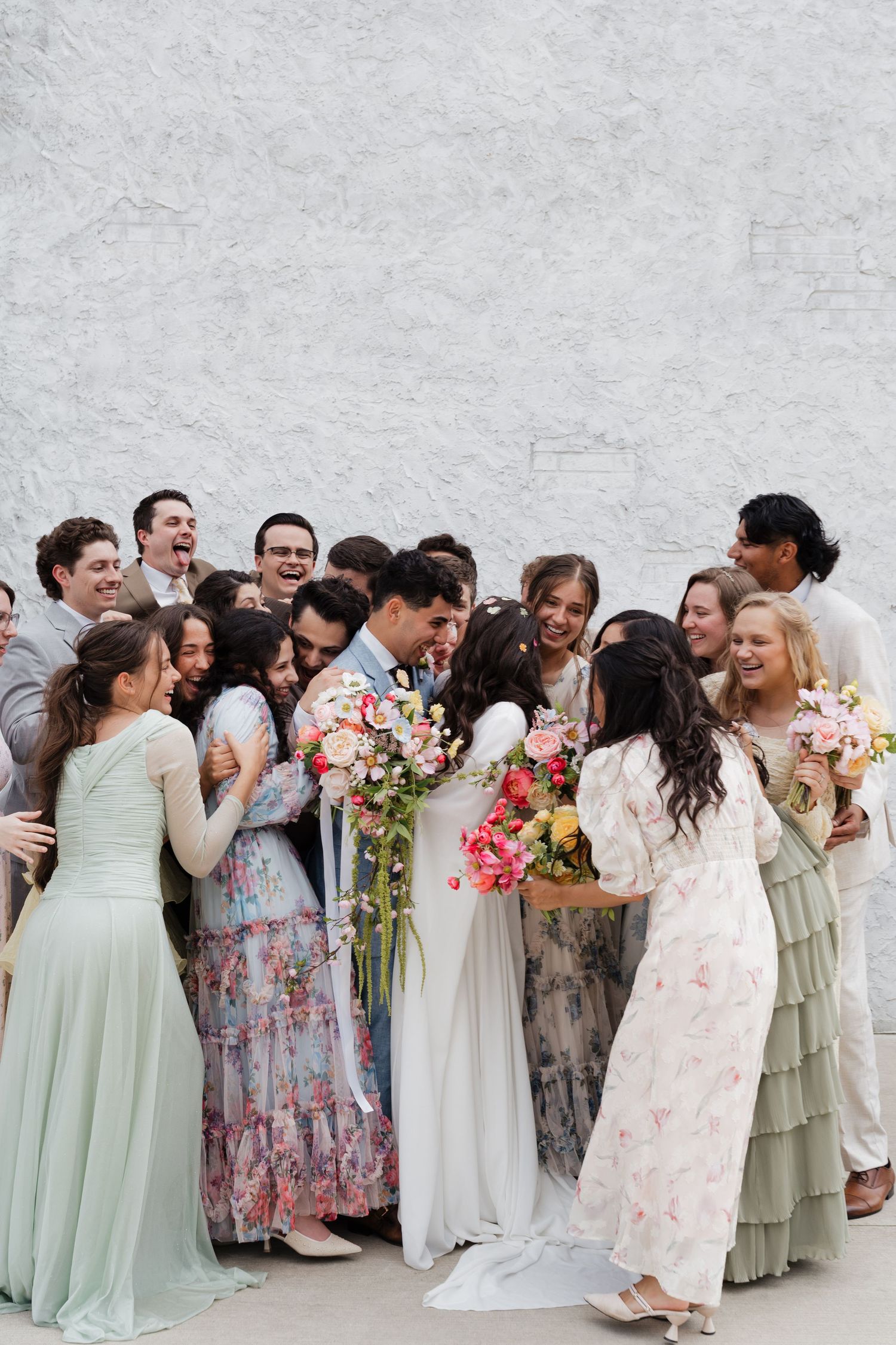 A joyful wedding party surrounds the bride and groom as they kiss, with colorful floral bouquets and pastel dresses.